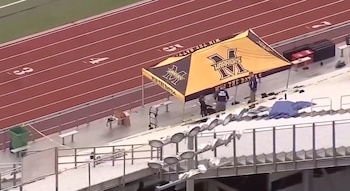 Vista aérea de un estadio deportivo con pista de atletismo. Una carpa amarilla y negra con "Warriors" y "POLICE" visible. Dos figuras y una lona azul en el suelo junto a gradas