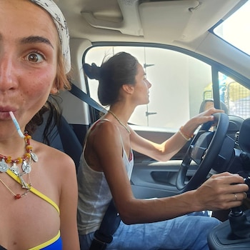 Una mujer con bandana y collar bebe con un popote junto a otra mujer que conduce un vehículo