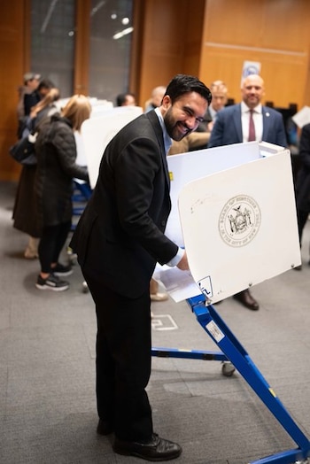 Un hombre de traje negro y camisa azul vota en una cabina electoral blanca con el escudo de Nueva York en una sala con otras personas