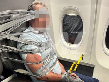 Un hombre sentado en un avión junto a una ventanilla aparece sujetado al respaldo de su asiento y en el torso con cinta adhesiva gris