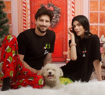 Fiorella Rodríguez e Ivan Micol, con su perro blanco, sentados en un tapete peludo frente a un fondo navideño rojo. Visten pijamas y camisetas del Grinch