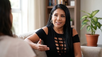 Tula Rodríguez, uma mulher de cabelos escuros e blusa preta com cinto, está sentada em um sofá, movendo-se enquanto fala com alguém fora da câmera.
