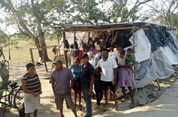 Un grupo de siete personas, incluidos cinco adultos y dos niños, posan en un área rural frente a un refugio hecho con postes de madera, lonas y plásticos