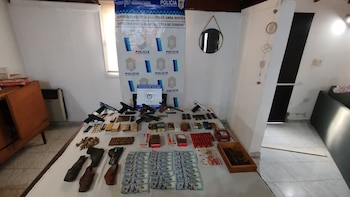 Una mesa con armas de fuego, fundas, cajas de municiones, cartuchos y billetes exhibidos frente a un cartel de la Policía de Buenos Aires