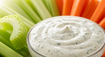Primer plano de un recipiente de vidrio con aderezo ranch blanco y hierbas, rodeado de palitos de apio verdes y palitos de zanahoria anaranjados.
