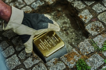 Primer plano de una mano enguantada sobre un 'Stolperstein' de latón dorado incrustado en un pavimento de adoquines grises y tierra