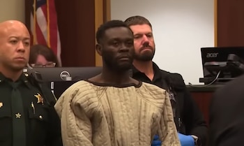 Hombre con barba y bata acolchada beige es escoltado por policías en un tribunal. Se ven monitores de computadora y una bandera estadounidense en el fondo