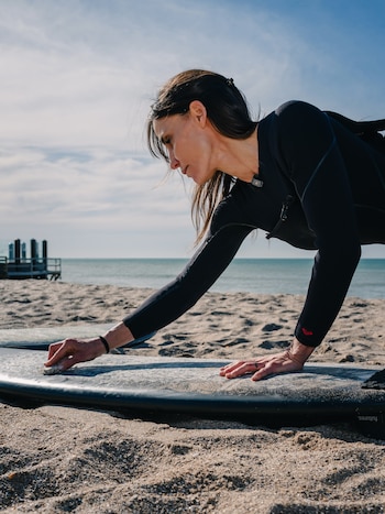 Una mujer con traje de neopreno negro aplica cera sobre una tabla de surf apoyada en la arena de la playa