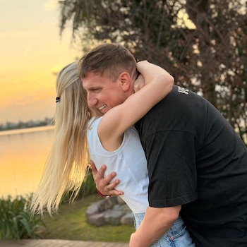 Maxi López abraza sonriente a Daniela Christiansson de espaldas, con cabello rubio y largo, contra un fondo de atardecer dorado sobre un lago