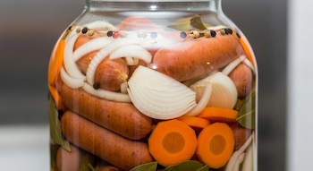 Frasco de vidrio grande con salchichas, rodajas de cebolla y zanahoria sumergidas en escabeche transparente, fondo neutro.