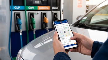 Manos sostienen un teléfono móvil con un mapa digital frente al capó de un automóvil y un surtidor de combustible con varias mangueras.