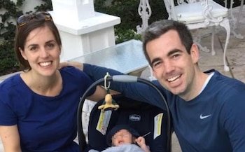 Una mujer y un hombre sonríen sentados junto a un portabebés con un bebé en un patio exterior con sillas blancas
