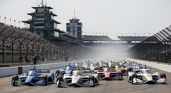 Vista del pelotón de coches de carrera IndyCar en la pista al inicio de la Indy 500, con tribunas llenas y la torre de control de fondo.