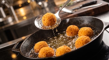 Bolas de papa empanadas doradas en aceite dentro de una sartén de hierro y una espumadera metálica levantando una.