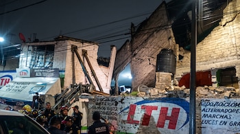 Elementos de la policía y bomberos frente a una edificación de ladrillo con estructuras derrumbadas y maquinaria pesada durante la noche.
