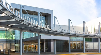 Vista frontal del Hospital Comunitario Martin Luther King Jr. con entrada cubierta, pilares, fachada de vidrio y paneles grises, y paisaje alrededor