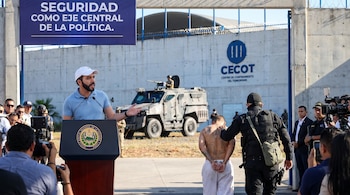 Vista del presidente Nayib Bukele hablando en un podio con el emblema de El Salvador, frente al CECOT, un vehículo blindado y un detenido esposado siendo escoltado.