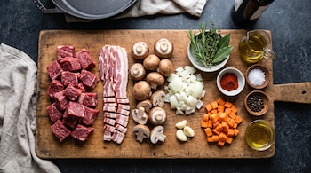 Carne picada en cubos, lonchas de panceta, champiñones, cebolla, zanahoria, dientes de ajo y especias sobre una tabla de cortar de madera.