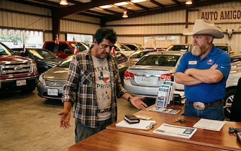 Un hombre vestido con camisa a cuadros gesticula frente a un vendedor de autos con sombrero de vaquero y barba en un concesionario de Texas con varios vehículos