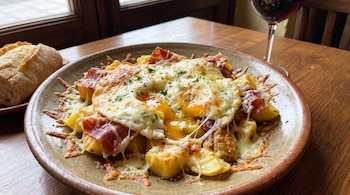 Un plato de huevos fritos con papas, jamón, queso fundido y perejil picado, junto a un trozo de pan y una copa de vino tinto en una mesa.