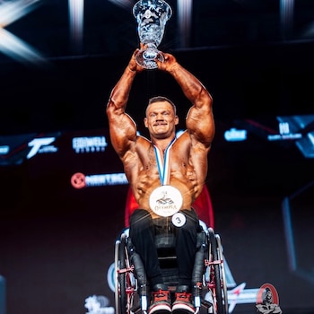 Un hombre musculoso sentado en una silla de ruedas sostiene un trofeo de cristal sobre su cabeza mientras luce una medalla en el pecho