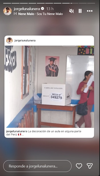 Interior de un aula convertida en cabina de votación con una mesa y una caja electoral. En la pared, un póster de Jorge Luna graduado. Una mujer camina