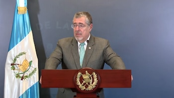 Hombre con gafas y barba canosa, vestido con traje y corbata, hablando desde un podio de madera con emblema dorado. A su izquierda, la bandera de Guatemala