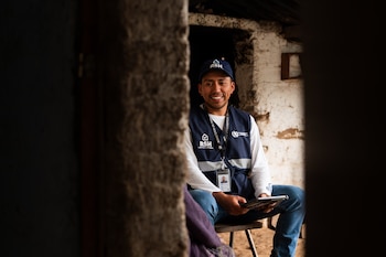 Un hombre joven con gorra y chaleco de Registro Social de Hogares de Guatemala se sienta y sonríe, sostiene una tableta, con credencial y fondo de pared rústica