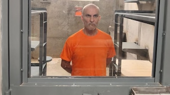 Un hombre rapado con camiseta naranja está dentro de una celda de prisión, visto a través del cristal de la puerta