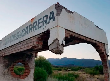 Pórtico de ladrillo y yeso blanco desmoronándose con la palabra 'CARRERA' pintada, rodeado de vegetación árida y montañas lejanas bajo un cielo claro