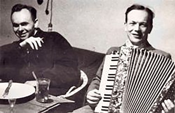 Foto en blanco y negro de dos hombres; uno toca un acordeón y el otro, sonriente, está sentado a una mesa con una bebida