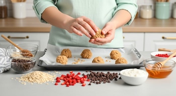Manos forman bolitas de masa en charola con papel encerado. Avena, chispas de chocolate, frutos rojos y miel en recipientes sobre encimera de cocina.