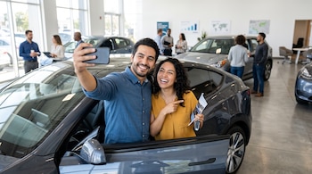Un hombre y una mujer sonríen mientras se toman una foto con un teléfono móvil junto a un coche gris en un concesionario.