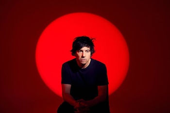 Hombre de pelo oscuro y camiseta negra sentado frente a un fondo rojo oscuro, con un círculo de luz roja intensa detrás de su cabeza. Manos entrelazadas