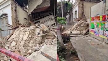 Escombros de una pared derrumbada, incluyendo ladrillos, madera y concreto, en una calle. Edificio con puerta arqueada y ventanas, cinta de "Peligro" roja