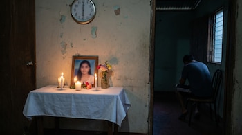 En una habitación con poca luz, un altar con la fotografía de una joven, velas encendidas y flores. Un hombre de espaldas sentado en una silla.