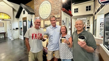 Cuatro personas, tres hombres y una mujer, sonríen a la cámara sosteniendo tazas de café dentro de un museo con réplicas de casas y edificios de ladrillo de fondo