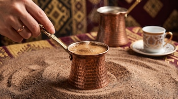 Una mano sostiene una cafetera de cobre con café hirviendo sobre arena, junto a otra cafetera y una taza de porcelana con plato.