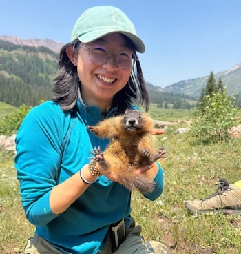 Mujer joven con gafas y gorra verde sonríe, sosteniendo una marmota de pelaje marrón. Detrás hay montañas, vegetación y cielo azul claro. Una bota de senderismo