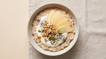 Un bowl de cerámica con avena, yogur, rodajas de manzana, nueces, semillas de chía, semillas de calabaza y semillas de girasol. Fondo beige y textil.