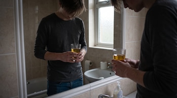 Un hombre con la cabeza inclinada sostiene un vaso con líquido amarillento frente al espejo de un baño.