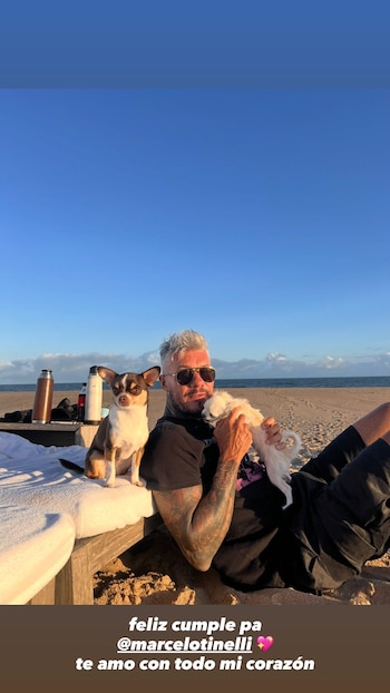 Un hombre con cabello gris y gafas de sol, recostado en la arena de una playa, sostiene un perro blanco mientras otro perro tricolor se sienta a su lado