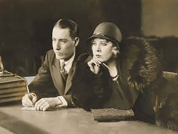 Imagen monocromática de Hunter Metts escribiendo en una mesa, con una mujer a su lado, vestida con sombrero y abrigo de piel, observando