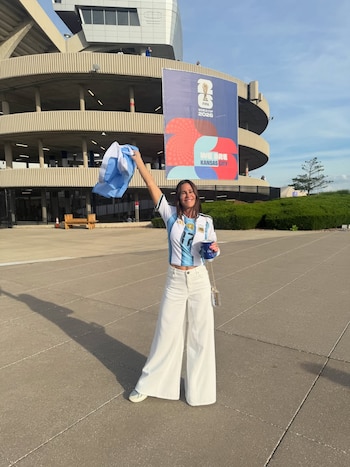 Una mujer sonriente, Carolina Baldini, vestida con una camiseta de fútbol argentina y pantalones blancos, sostiene una bandera de Argentina fuera de un estadio