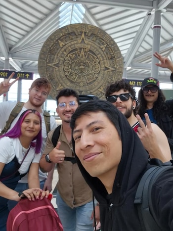 Seis personas posan para una selfie delante de una réplica circular grande del Calendario Azteca. Se observa un techo de vigas metálicas y carteles