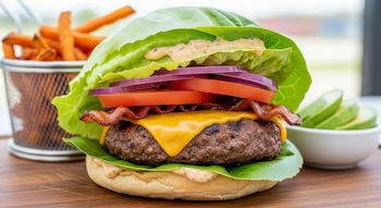 Primer plano de una hamburguesa envuelta en lechuga con carne, queso, tocino, tomate, cebolla morada; batatas fritas y aguacate al fondo.