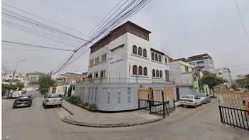 Vista panorámica del Colegio Salvador, un edificio blanco con techo de madera y ventanas arqueadas, rodeado por una verja y calles con coches estacionados bajo un cielo gris