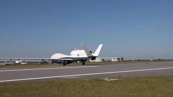 Primer plano de un Northrop Grumman RQ-4 Global Hawk blanco y gris en una pista de asfalto. El dron tiene alas largas y cola en V. Cielo azul claro y edificios al fondo