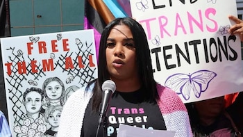 Mujer con cabello oscuro y blusa negra "Trans Queer" habla por un micrófono frente a carteles de protesta que piden "FREE THEM ALL" y el fin de las detenciones trans