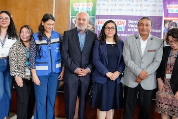 Siete personas, cuatro mujeres y tres hombres, de pie frente a un banner con mensajes sobre vacunación y logos de OPS y UNICEF, vestidas con ropa formal y casual